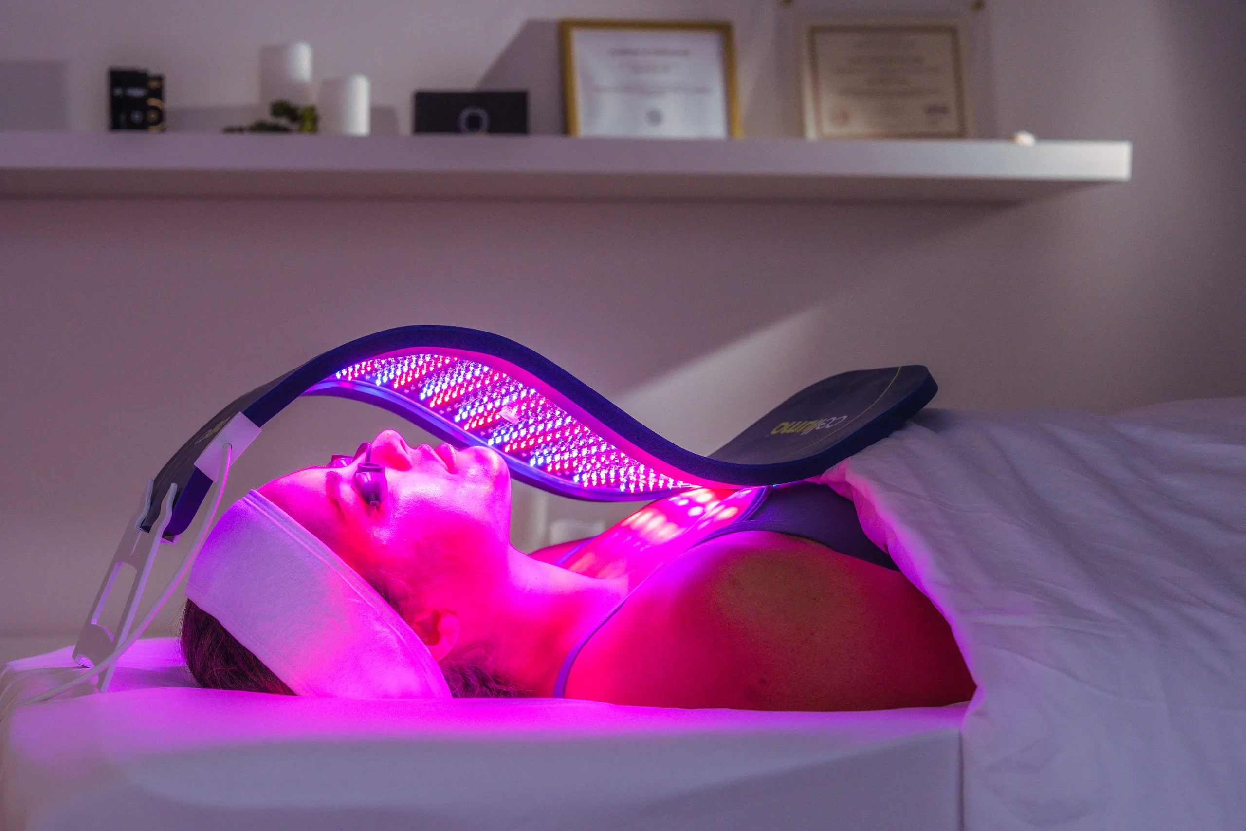 Mujer recibe un tratamiento facial con terapia de luz LED roja y violeta mediante un dispositivo especializado, con los ojos cerrados y recostada en una camilla.